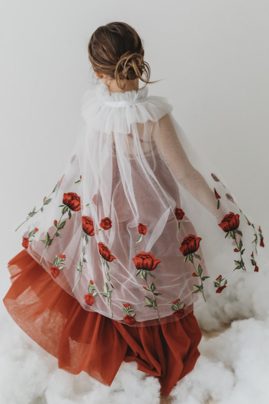 A young girl is wearing a red dress with a white tulle cape adorned with embroidered red roses. The floral details of her ensemble are striking. The shades of red and green are the perfect addition to our Red Rose Princess Collection.