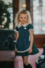 A young girl stands indoors wearing a dark teal green dress with white lace trim, a matching plaid blue and green apron, puff sleeves and a white collar. The woven cotton is perfect for spooky season.