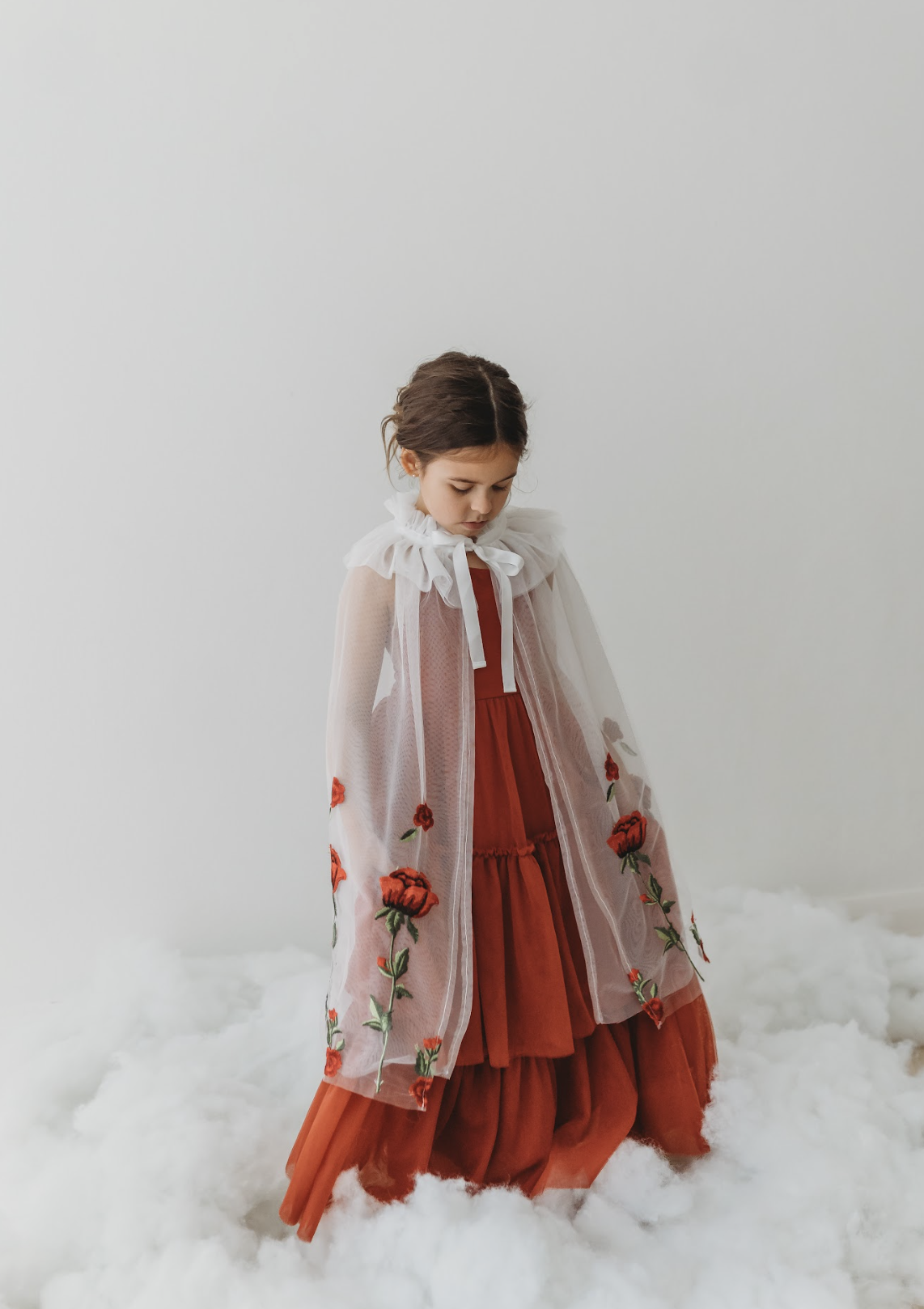A young girl is wearing a red dress with a white tulle cape adorned with embroidered red roses. The floral details of her ensemble are striking. The shades of red and green are the perfect addition to our Red Rose Princess Collection.