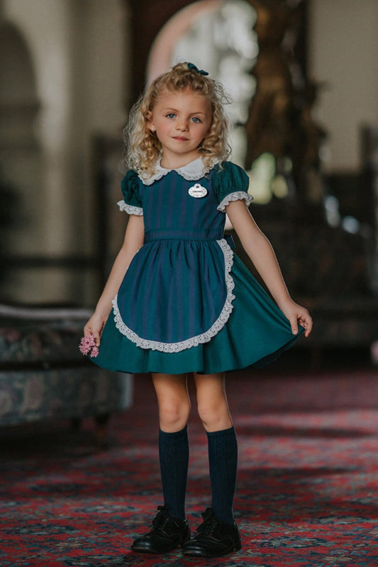 A young girl stands indoors wearing a dark teal green dress with white lace trim, a matching plaid blue and green apron, puff sleeves and a white collar. The woven cotton is perfect for spooky season.