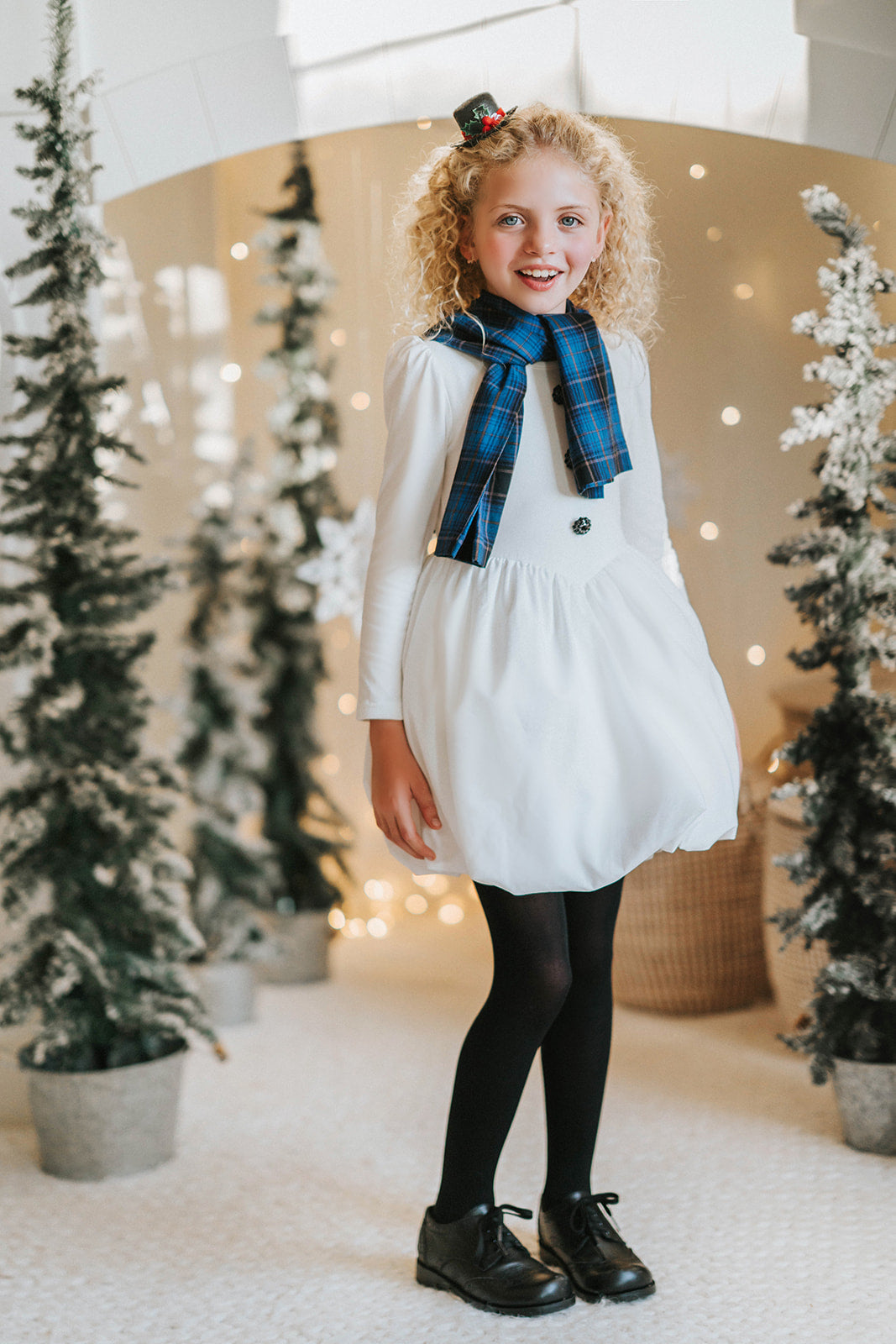 Girl wearing white holiday dress with bubble hem, black tights, and a plaid scarf, styled as a snowman outfit for Christmas photos, winter events, or holiday parties.
Child in snowman-inspired winter dress with black leggings, puff hem skirt, and blue plaid scarf, perfect for Christmas card photos and seasonal festivities.
