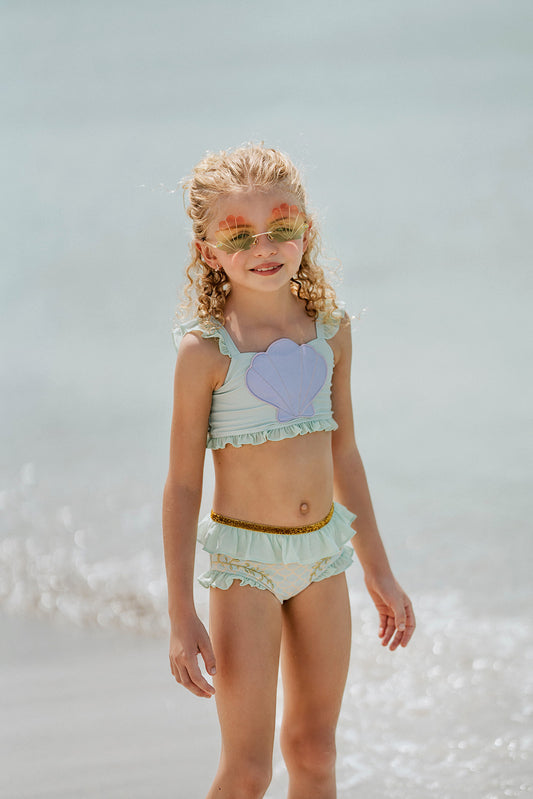 Girl wearing mint green mermaid swimsuit with purple seashell top and gold waistband, standing at the shoreline in fun sunglasses
Mermaid-themed ruffle bikini for girls with shell applique and scale print bottoms, styled for beach photos and play
Child in seafoam green two-piece swimsuit with frilly straps and shell detail, posing on sandy beach in sunshine
