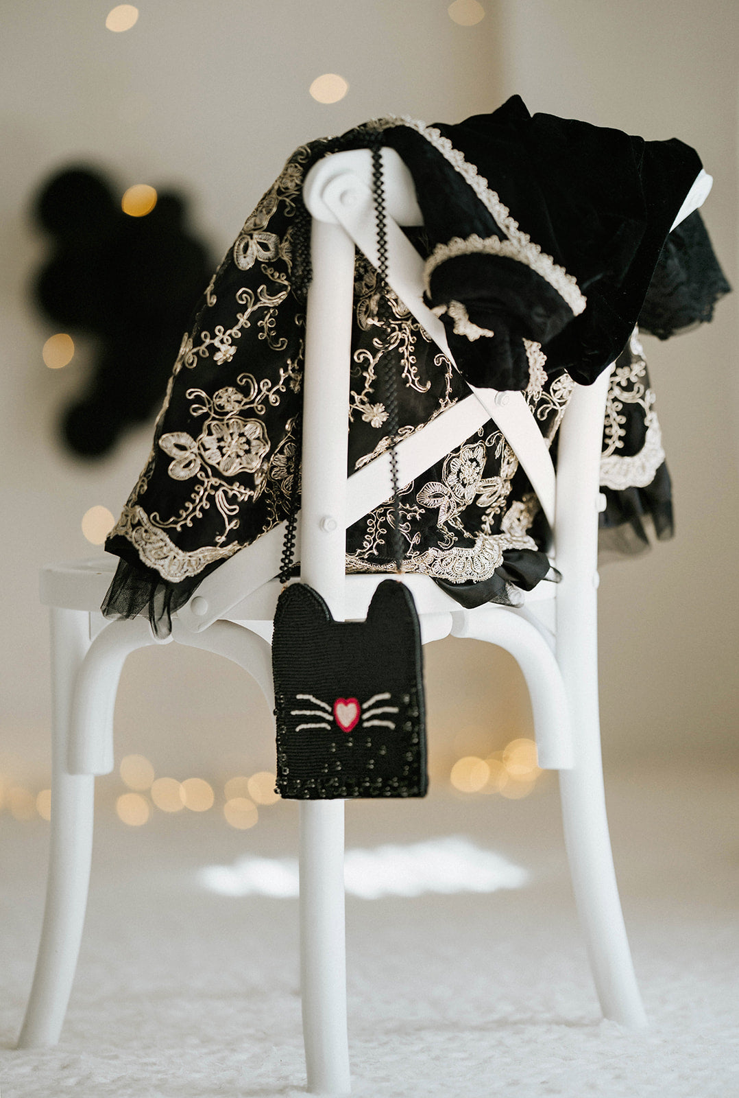 Black beaded cat purse with heart-shaped nose and whiskers, styled with lace-trimmed gothic dress on a white chair for Halloween trick-or-treat or costume party accessory.
Whimsical black cat crossbody bag for girls with beaded design, heart nose, and whiskers, styled next to embroidered Halloween dress for spooky season outfit inspiration.
Close-up of handmade cat-shaped purse featuring sparkly black seed beads and pink heart nose, hanging from white chair for Halloween, gothic, or dress-up styling.