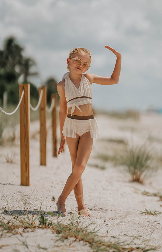 Girl wearing cream two-piece wrap swimsuit with halter top and flowy side skirt, posing confidently on a sandy beach
Boho-inspired cream girls swimsuit with wrap detail and side draping, styled for beach photos or summer vacation
Toddler to tween girl in goddess-style cream bathing suit with halter neckline and high-waisted bottoms, standing on the shore