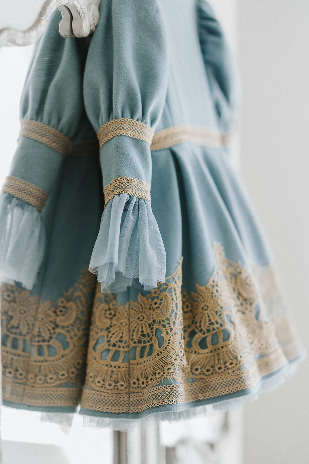 Elegant heirloom girls dress in dusty blue with gold lace trim and tulle layers, hanging on ornate vintage-style mirror
Long sleeve blue and gold embroidered holiday dress for girls, featuring pearl buttons and layered tulle hem
Girls vintage-style formal gown in soft blue with gold lace and flounce sleeves, ideal for portraits or weddings
Classic blue heirloom dress for toddlers and girls with ornate embroidery and pearl detailing, styled for a timeless photo shoot