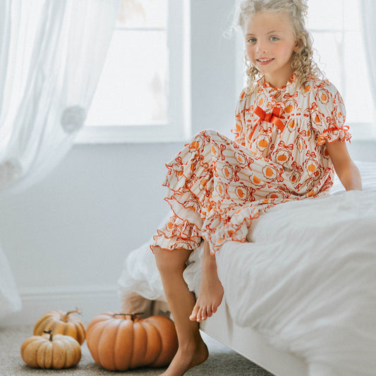 Young girl in a floral dress sitting on a bed with pumpkins beside her