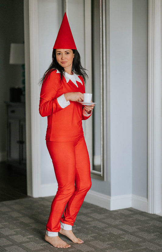 A young child dressed in Unisex Elf Pajamas with a red pointy hat. The perfect Christmas photo outfit. The fabric is bright red and buttery soft. The top is long sleeved with white cuffs and a white collar that has large triangle points similar to a clown. Long pants with white cuffs.