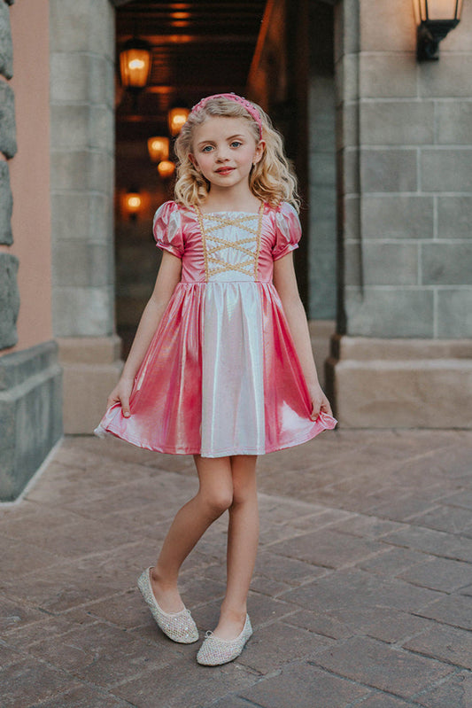 A young girls is wearing a metallic Sleeping Beauty Twirl dress. The pink and white knee length dress has a criss cross gold ribbon across the bodice. The ballroom puff sleeves add elegance.