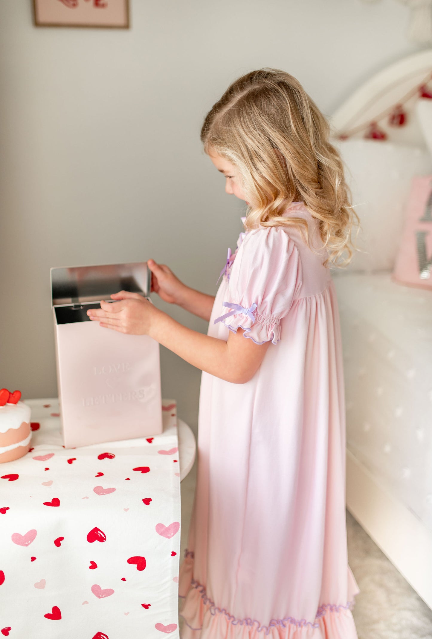 A young girl is wearing The Original Clara Nutcracker Gown in Blush, featuring blush pink ruffles and a lavender bow. The buttery soft fabric is cozy and sensory friendly. The puff sleeves with ribbon detail and the ruffles at the hem add a traditional classic look.