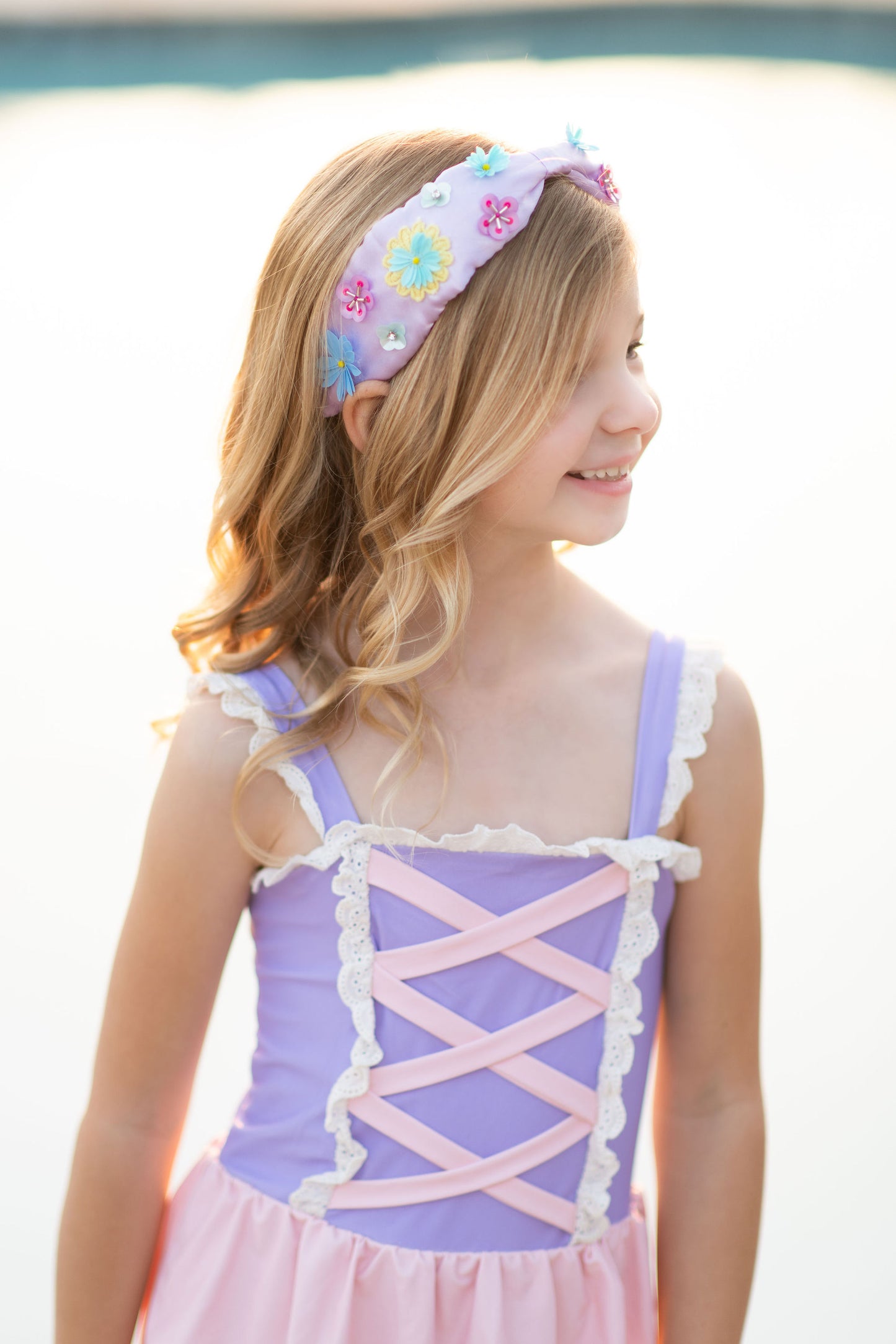 A young girl stands on a beach wearing a pastel pink and purple Rapunzel One Piece Swimsuit, adorned with crisscross and lace detail. The bodice is lavender and skirt is light pink. The swimsuit is SPF 50 for protection from the sun.