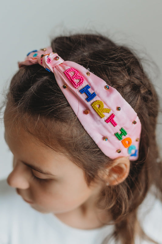 The light pink satin headband has "Happy Birthday" spelled out in colorful beading. Each letter is a bight color of pink, blue, yellow, green or orange. There are multicolor rhinestones as well. The center has a small tied knot. This hair accessory is designed for all day comfort.