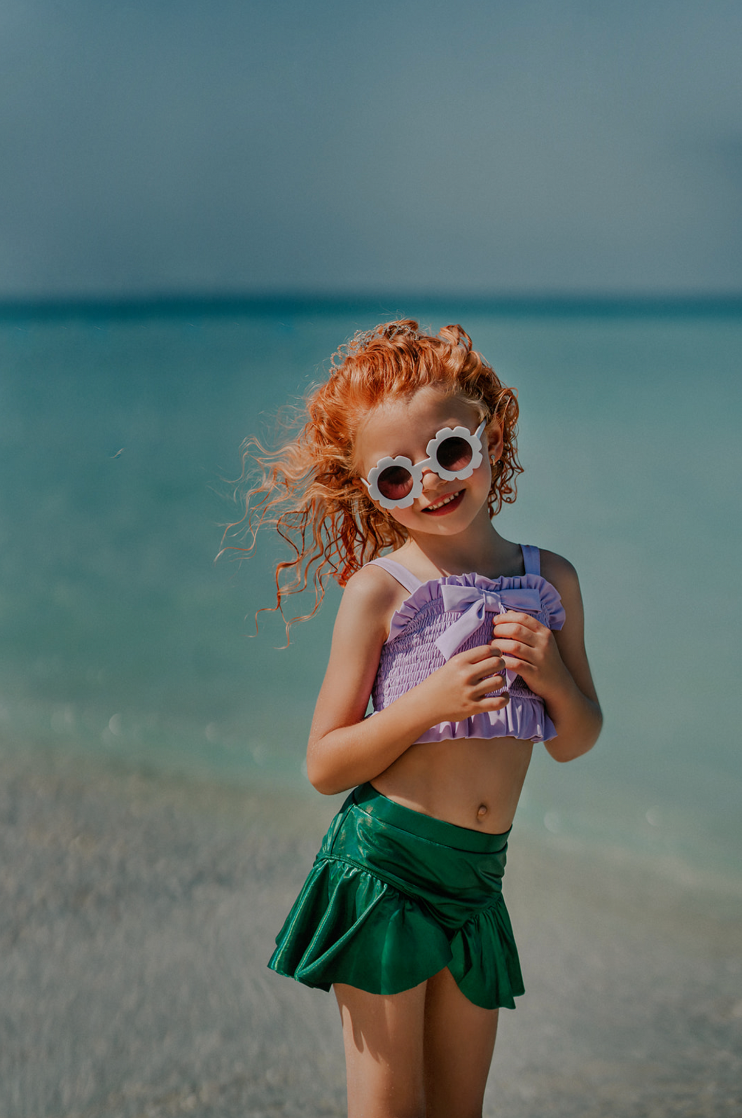 A young girl playing in the water is wearing the Mermaid Tail 2pc Swim set. The top is light purple with ruched bodice and ruffles. There is a bow in the center. The metallic green bottom is cut to resemble a mermaid and is longer on the sides.