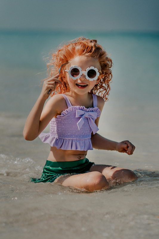A young girl playing in the water is wearing the Mermaid Tail 2pc Swim set. The top is light purple with ruched bodice and ruffles. There is a bow in the center. The metallic green bottom is cut to resemble a mermaid and is longer on the sides.
