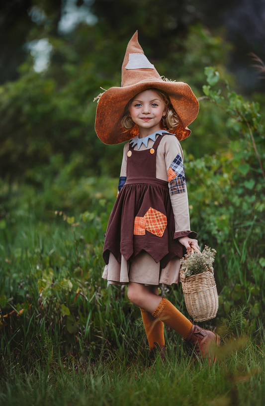 A young girl wears a tan long sleeve cotton Scarecrow dress. The dress has a jester triangle collar in grey. There is an overall brown apron with handkerchief bottom. There are orange and blue plaid patches on the sleeves and apron.