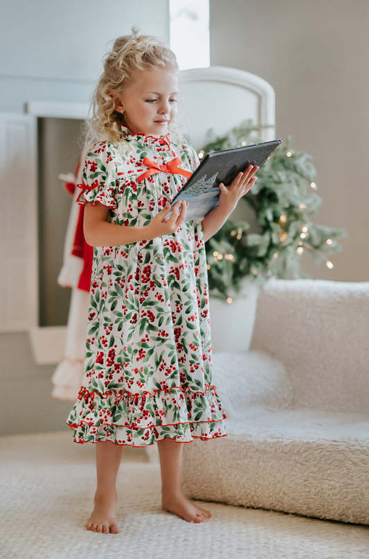 A young girl is wearing The Original Clara Nutcracker Gown in Winterberry, featuring winterberry print and a red bow. The buttery soft fabric is cozy and sensory friendly. The puff sleeves with ribbon detail and the ruffles at the hem add a traditional classic look.