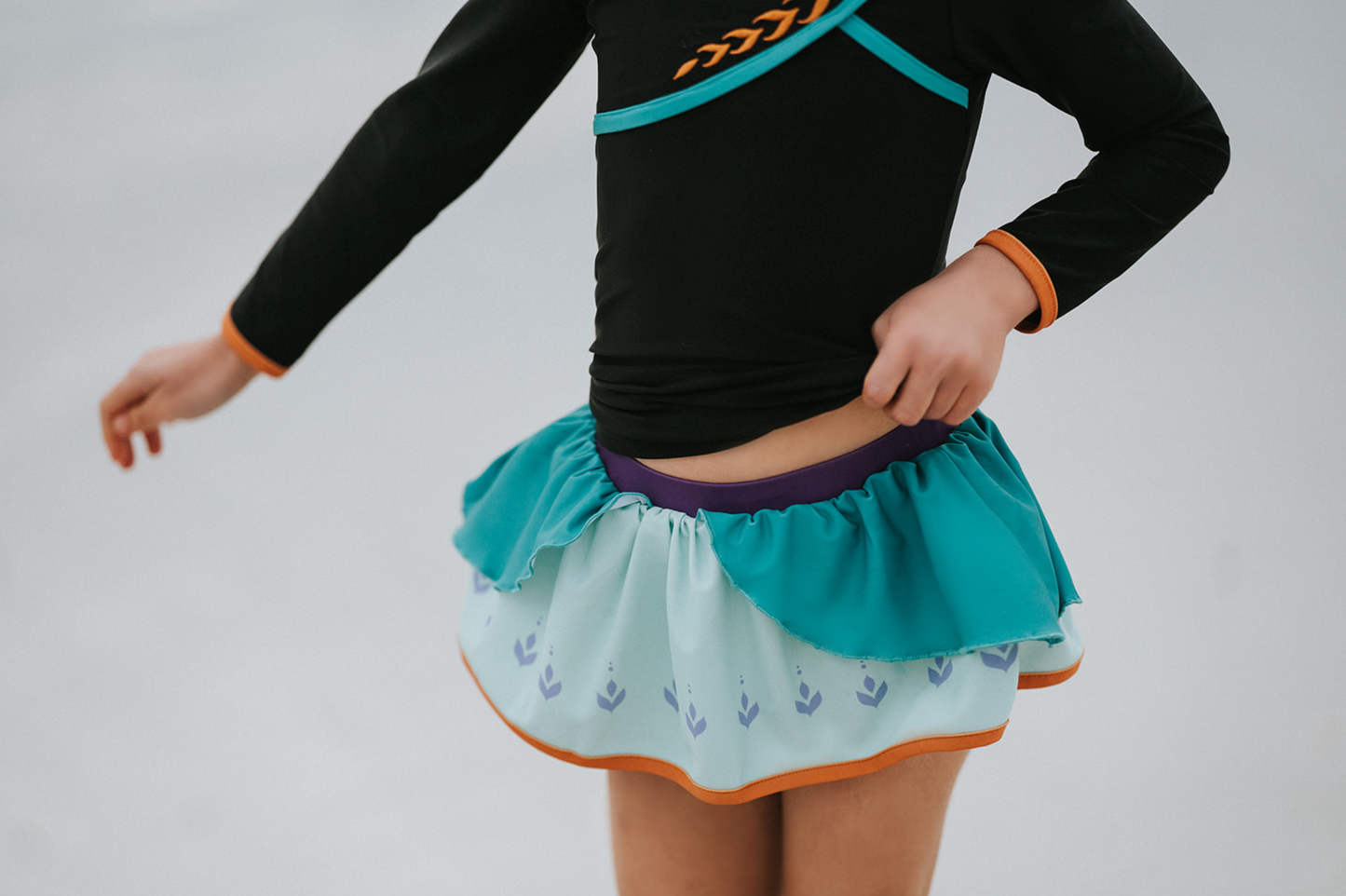 A young girl is wearing a 2 piece long sleeve rash guard swim set. The top is black with turquoise and goldenrod accents. The bottom has a turquoise ruffle over a mint teal skirt with goldenrod hem accent.