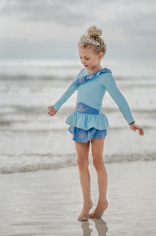 A young girl stands wearing a light blue 2 piece rash guard swim set. The long sleeve top has an asymetric cornflower blue ruffle with silver snow flake accents. The skirted bottom is cornflower blue and has silver snowflake.