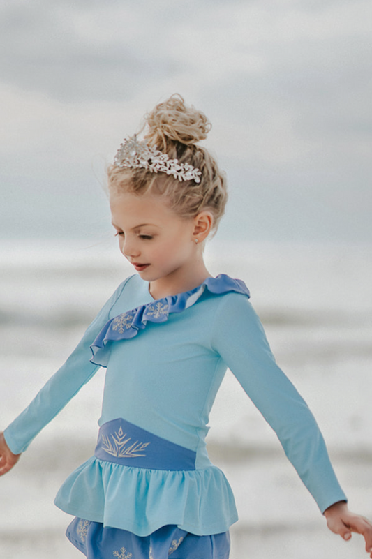 A young girl stands wearing a light blue 2 piece rash guard swim set. The long sleeve top has an asymetric cornflower blue ruffle with silver snow flake accents. The skirted bottom is cornflower blue and has silver snowflake.