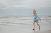 A young girl stands wearing a light blue 2 piece rash guard swim set. The long sleeve top has an asymmetric cornflower blue ruffle with silver snow flake accents. The skirted bottom is cornflower blue and has silver snowflake.