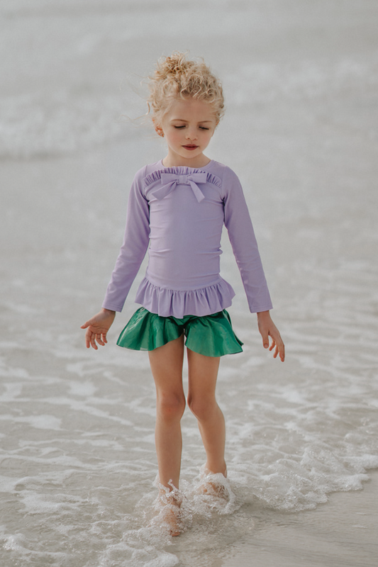 A young child walking on the shoreline at the beach is wearing a Mermaid Princess Rash Guard Swim Set with a long-sleeve purple top with a ruffle and bow at the chest. The modest bottom is metallic green and flares out at the sides to mimic a mermaid tail. The swim suit offers SPF 50 for protection from the sun.