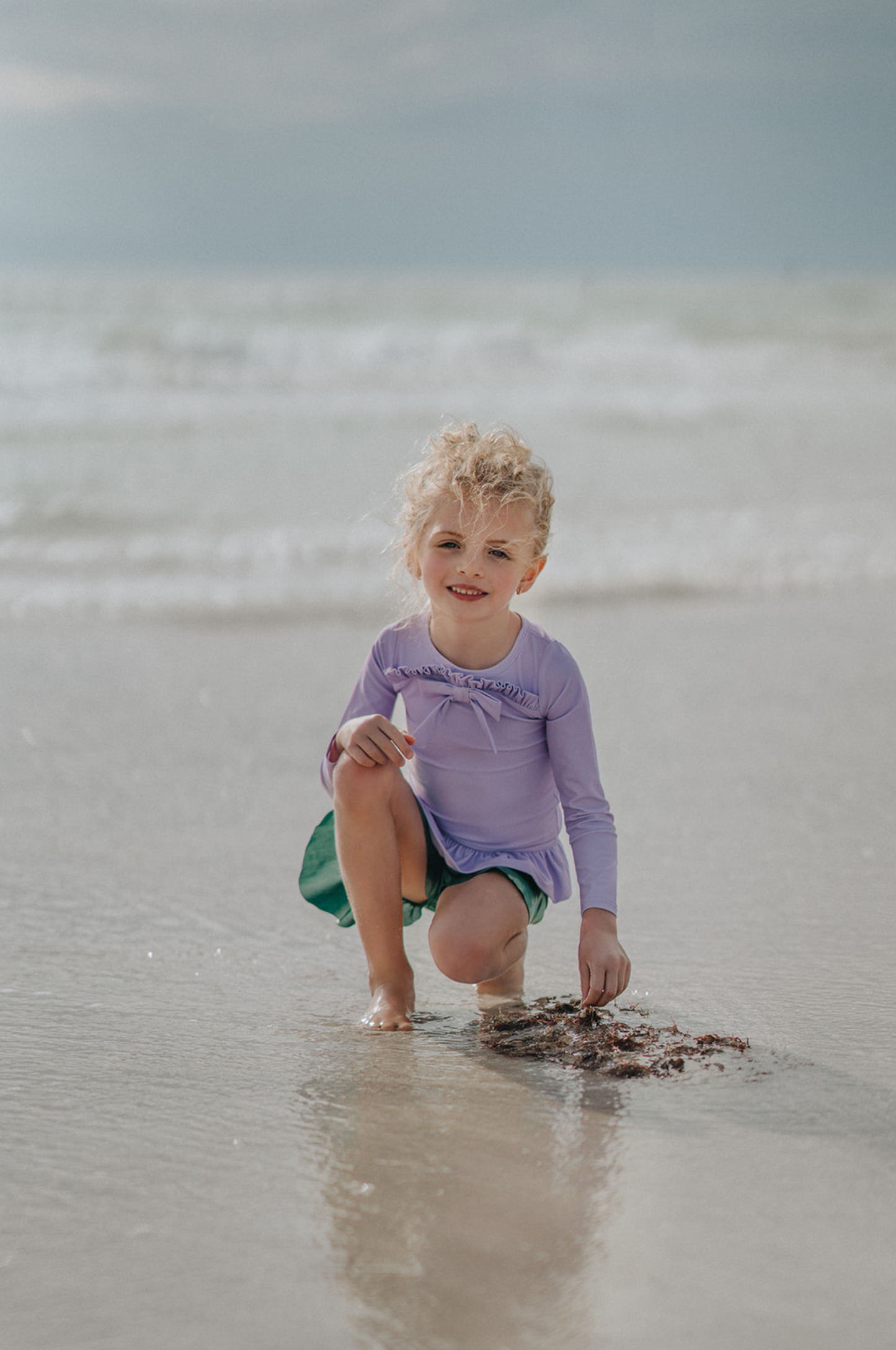 A young child walking on the shoreline at the beach is wearing a Mermaid Princess Rash Guard Swim Set with a long-sleeve purple top with a ruffle and bow at the chest. The modest bottom is metallic green and flares out at the sides to mimic a mermaid tail. The swim suit offers SPF 50 for protection from the sun.
