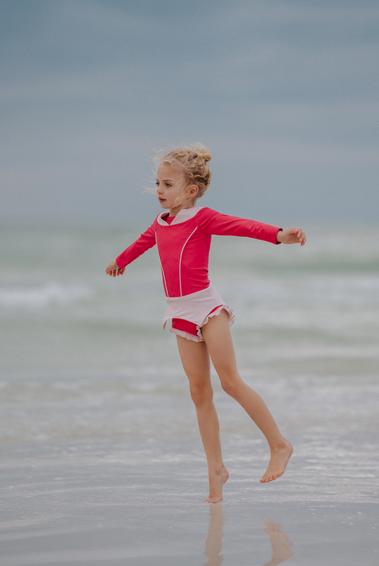 A young girl is wearing a pink 2 piece rashgaurd swim set. The Sleeping Beauty swim set has a pink collar that resembles a ballgown. The top has light pink piping and trim. The modest bottoms have a pink overlay and ruffles on the legs.