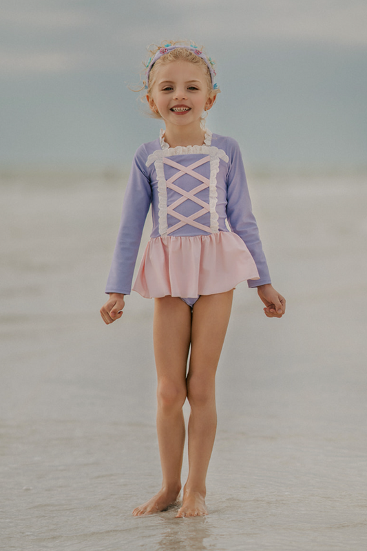 A young girl stands on a beach wearing a pastel pink and purple Rapunzel Rashguard Swimsuit, adorned with crisscross and lace detail. The bodice is lavender and skirt is light pink. The swimsuit is SPF 50 for protection from the sun.