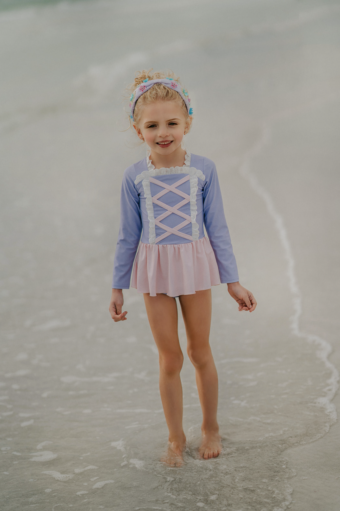 A young girl stands on a beach wearing a pastel pink and purple Rapunzel Rashguard Swimsuit, adorned with crisscross and lace detail. The bodice is lavender and skirt is light pink. The swimsuit is SPF 50 for protection from the sun.