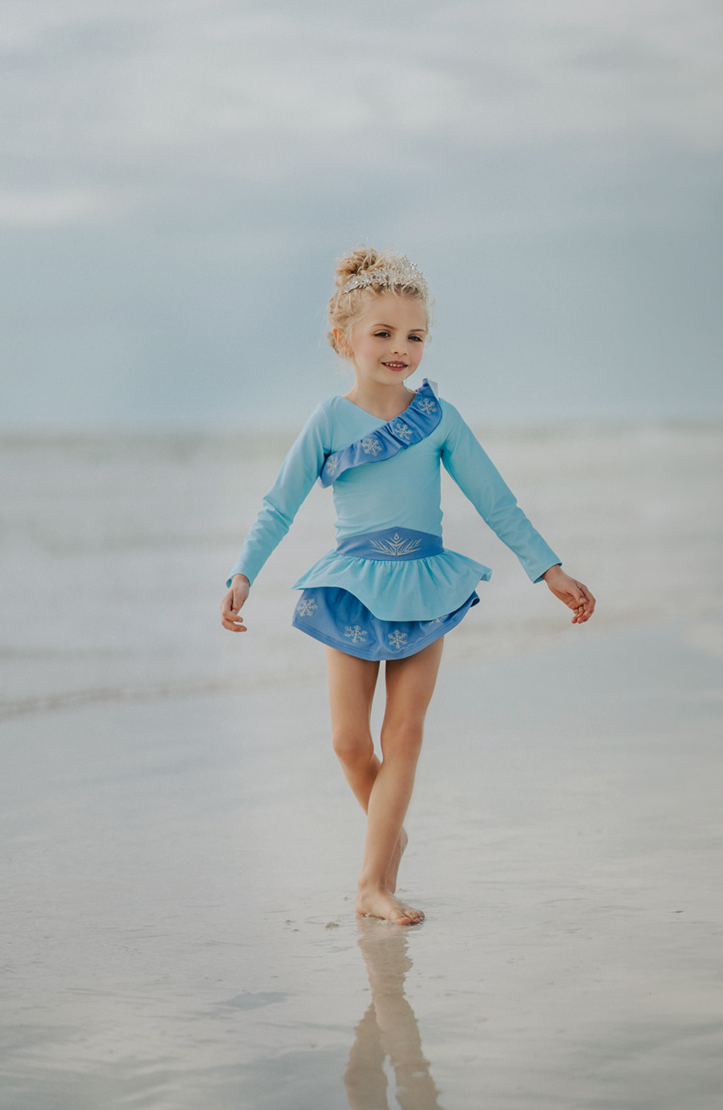 A young girl stands wearing a light blue 2 piece rash guard swim set. The long sleeve top has an asymmetric cornflower blue ruffle with silver snow flake accents. The skirted bottom is cornflower blue and has silver snowflake.