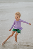 A young child walking on the shoreline at the beach is wearing a Mermaid Princess Rash Guard Swim Set with a long-sleeve purple top with a ruffle and bow at the chest. The modest bottom is metallic green and flares out at the sides to mimic a mermaid tail. The swim suit offers SPF 50 for protection from the sun.