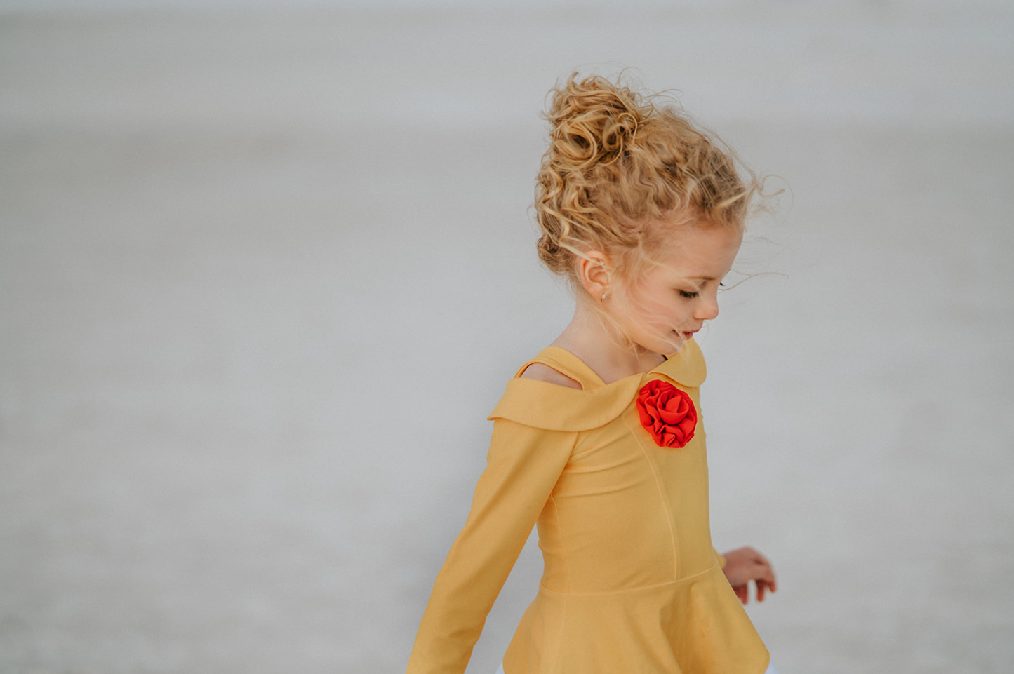 A young girl with curly blonde hair, wearing the Red Rose Rash Guard Swim Set featuring a yellow and white design with a red flower on the chest, stands on a beach with her feet in the shallow water. The sky is cloudy, and the ocean can be seen in the background.