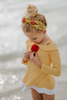 A young girl with curly blonde hair, wearing the Red Rose Rash Guard Swim Set featuring a yellow and white design with a red flower on the chest, stands on a beach with her feet in the shallow water. The sky is cloudy, and the ocean can be seen in the background.