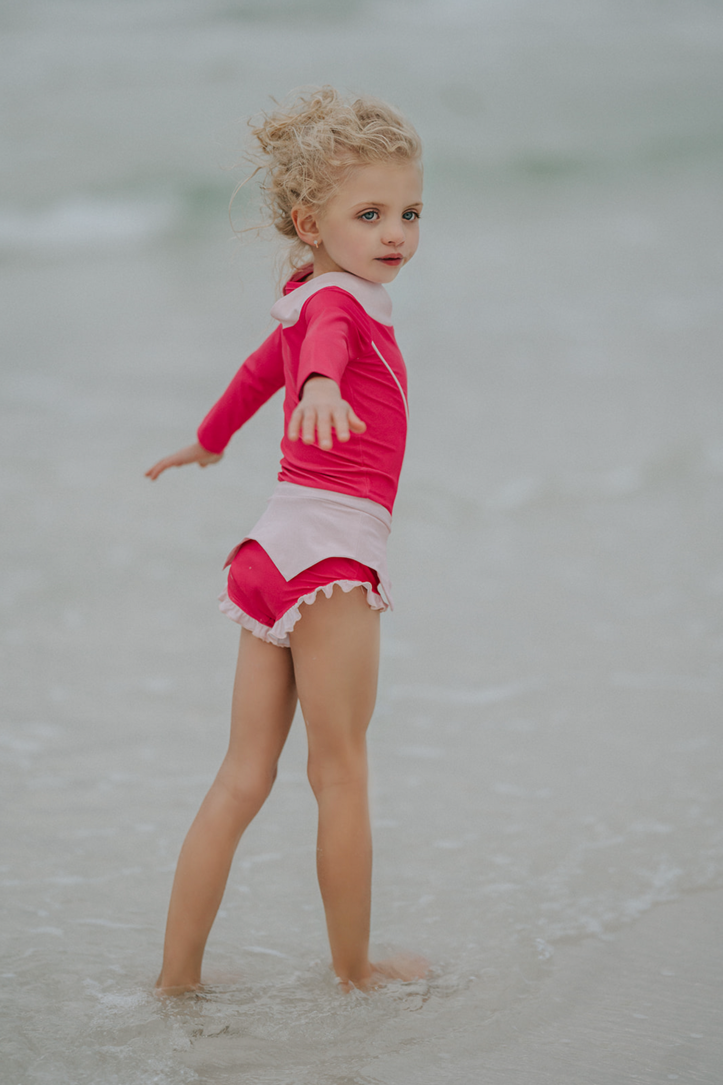 A young girl is wearing a pink 2 piece rashgaurd swim set. The Sleeping Beauty swim set has a pink collar that resembles a ballgown. The top has light pink piping and trim. The modest bottoms have a pink overlay and ruffles on the legs.