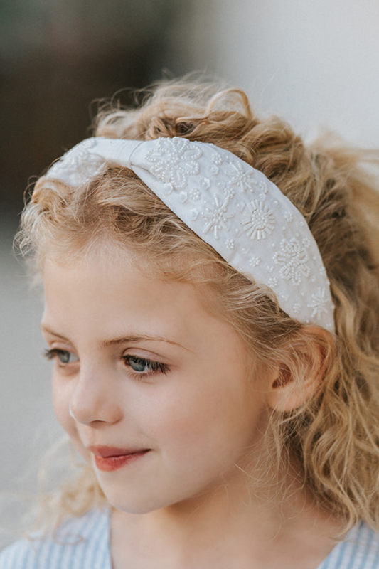 The Micro Beaded Floral Celebration Headband is white with small white white beads in various shapes. They are sewn in floral patterns and scattered to cover the headband. This hair accessory is designed for all day comfort.
