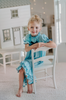 A young girl is wearing The Original Clara Nutcracker Gown in Robbins Egg Blue, featuring blue ruffles and a navy bow. The buttery soft fabric is cozy and sensory friendly. The puff sleeves with ribbon detail and the ruffles at the hem add a traditional classic look.