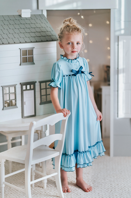 A young girl is wearing The Original Clara Nutcracker Gown in Robbins Egg Blue, featuring blue ruffles and a navy bow. The buttery soft fabric is cozy and sensory friendly. The puff sleeves with ribbon detail and the ruffles at the hem add a traditional classic look.