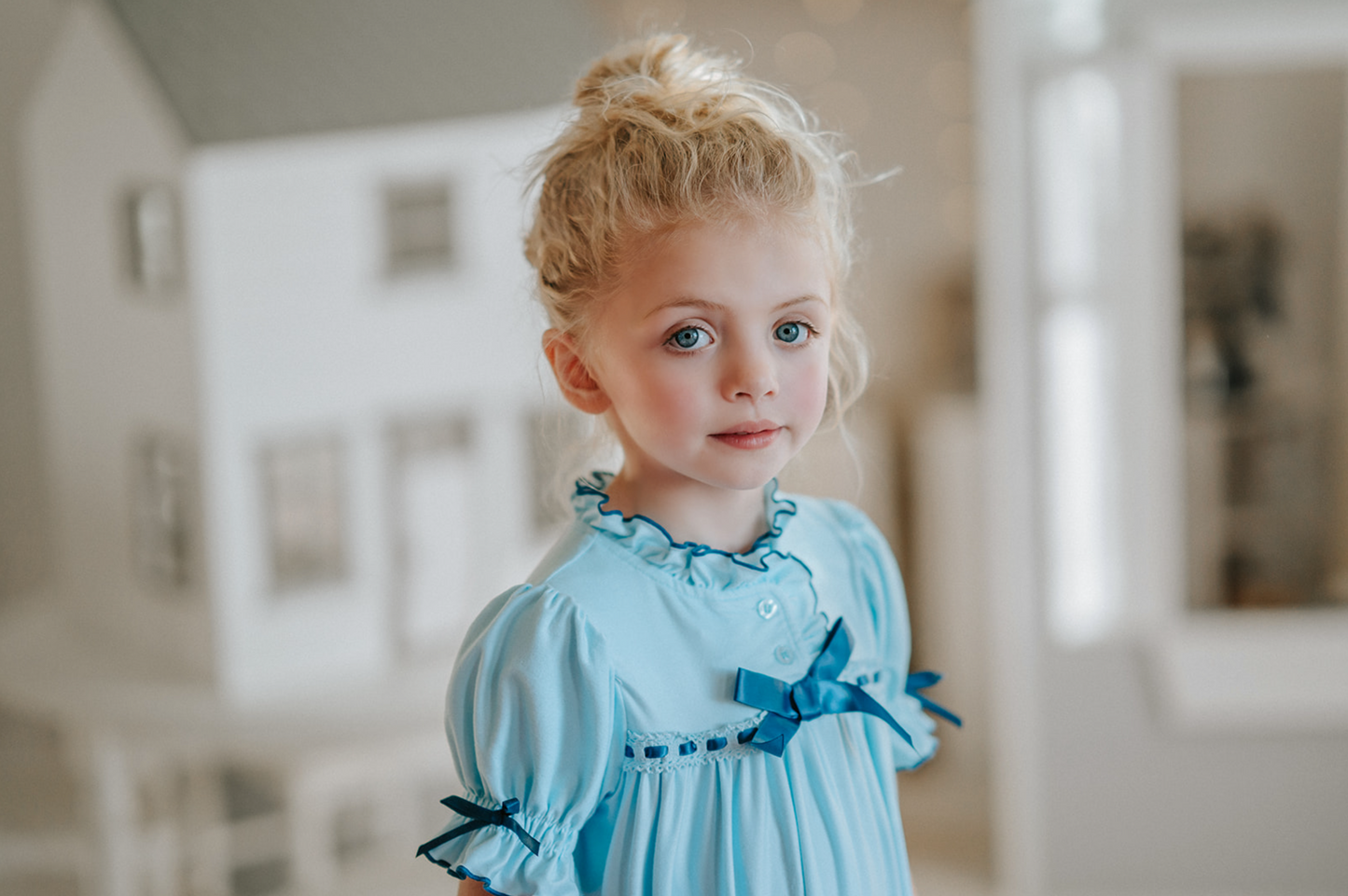 A young girl is wearing The Original Clara Nutcracker Gown in Robbins Egg Blue, featuring blue ruffles and a navy bow. The buttery soft fabric is cozy and sensory friendly. The puff sleeves with ribbon detail and the ruffles at the hem add a traditional classic look.