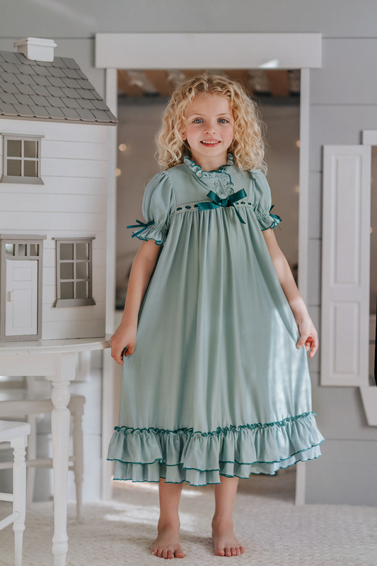 A young girl is wearing The Original Clara Nutcracker Gown in sage green, featuring green ruffles and a dark teal bow. The buttery soft fabric is cozy and sensory friendly. The puff sleeves with ribbon detail and the ruffles at the hem add a traditional classic look.