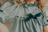 A young girl is wearing The Original Clara Nutcracker Gown in sage green, featuring green ruffles and a dark teal bow. The buttery soft fabric is cozy and sensory friendly. The puff sleeves with ribbon detail and the ruffles at the hem add a traditional classic look.