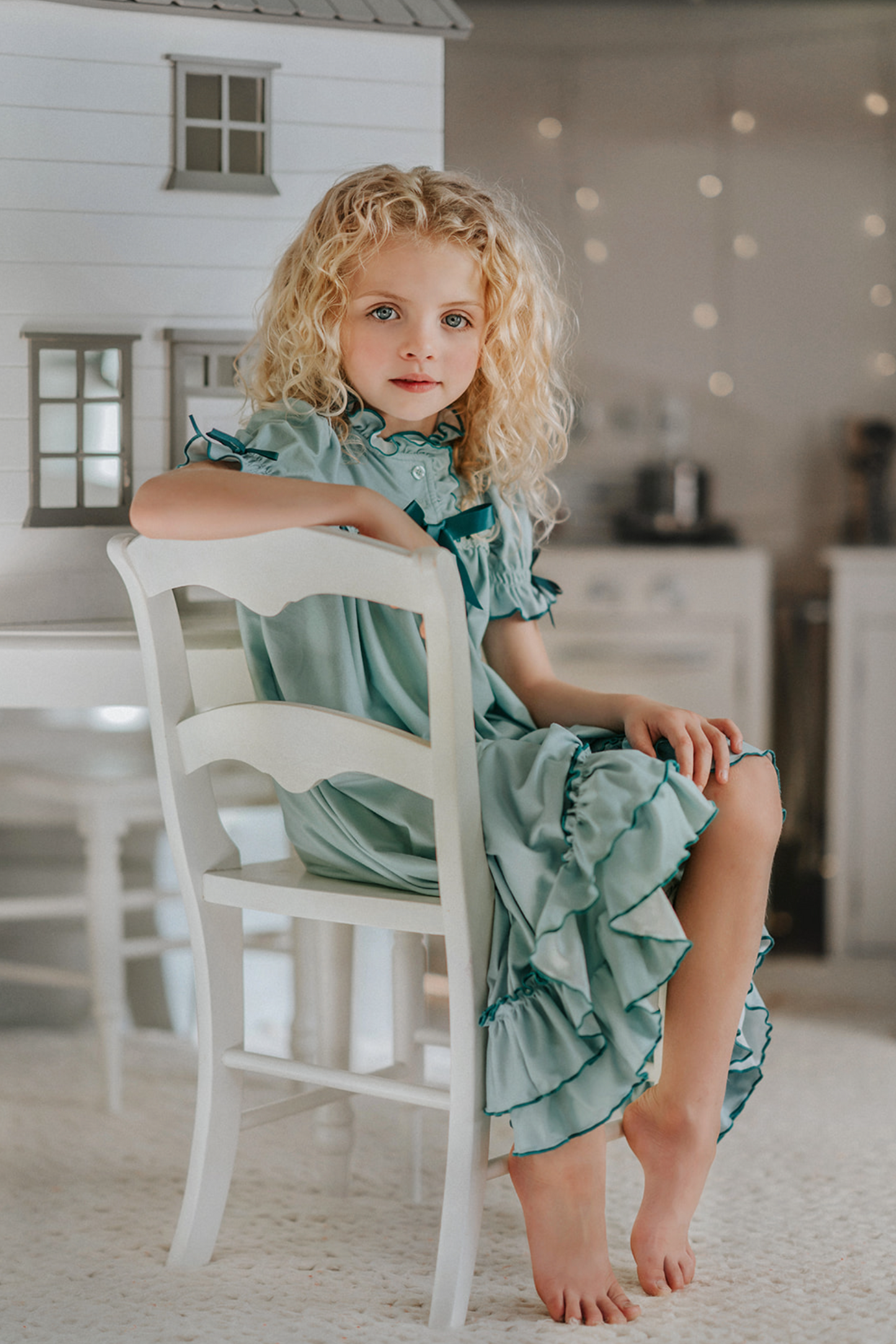 A young girl is wearing The Original Clara Nutcracker Gown in sage green, featuring green ruffles and a dark teal bow. The buttery soft fabric is cozy and sensory friendly. The puff sleeves with ribbon detail and the ruffles at the hem add a traditional classic look.