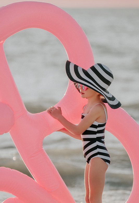 A young girl is wearing a black and white stripped one piece swim suit. The white spaghetti straps are adjustable. The black and white pattern is in a chevron style. A large floppy brimmed beach hat in matching black and white stripe is on her head.