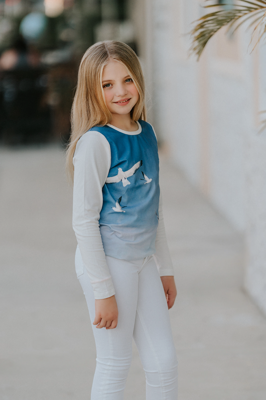 A young girl is wearing a stylish Seagull Soundtrack Shirt with a blue front panel featuring birds in flight, crafted from soft cotton jersey knit, paired with white leggings.