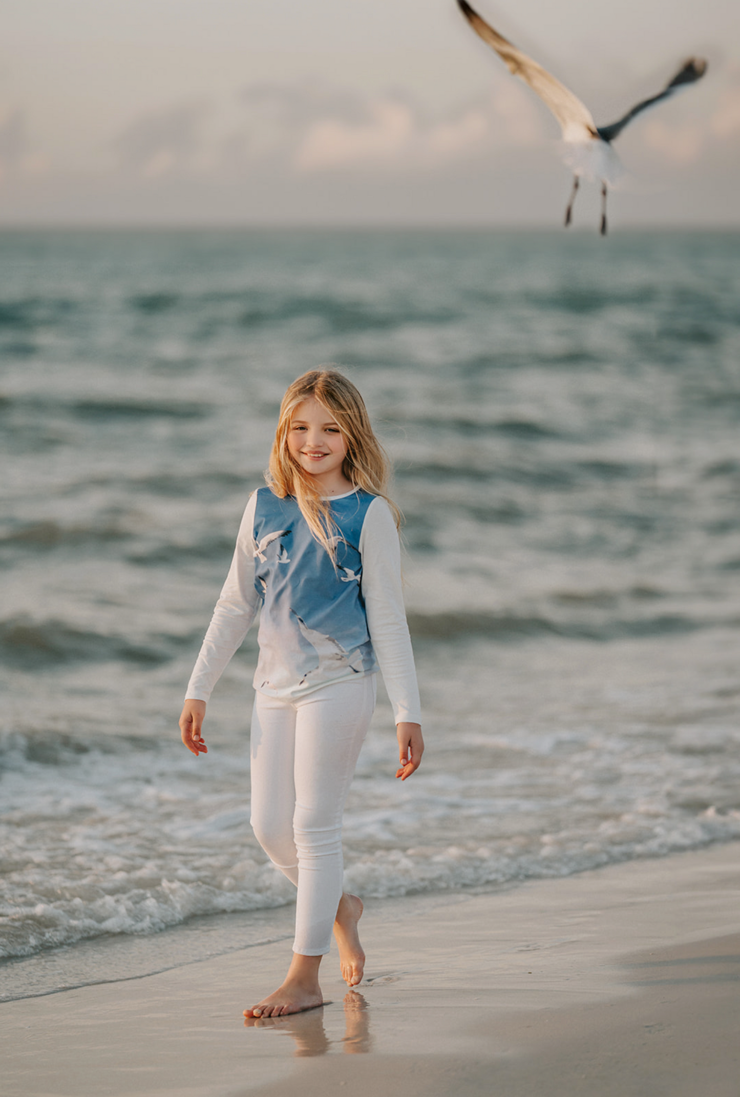A young girl is wearing a stylish Seagull Soundtrack Shirt with a blue front panel featuring birds in flight, crafted from soft cotton jersey knit, paired with white leggings.