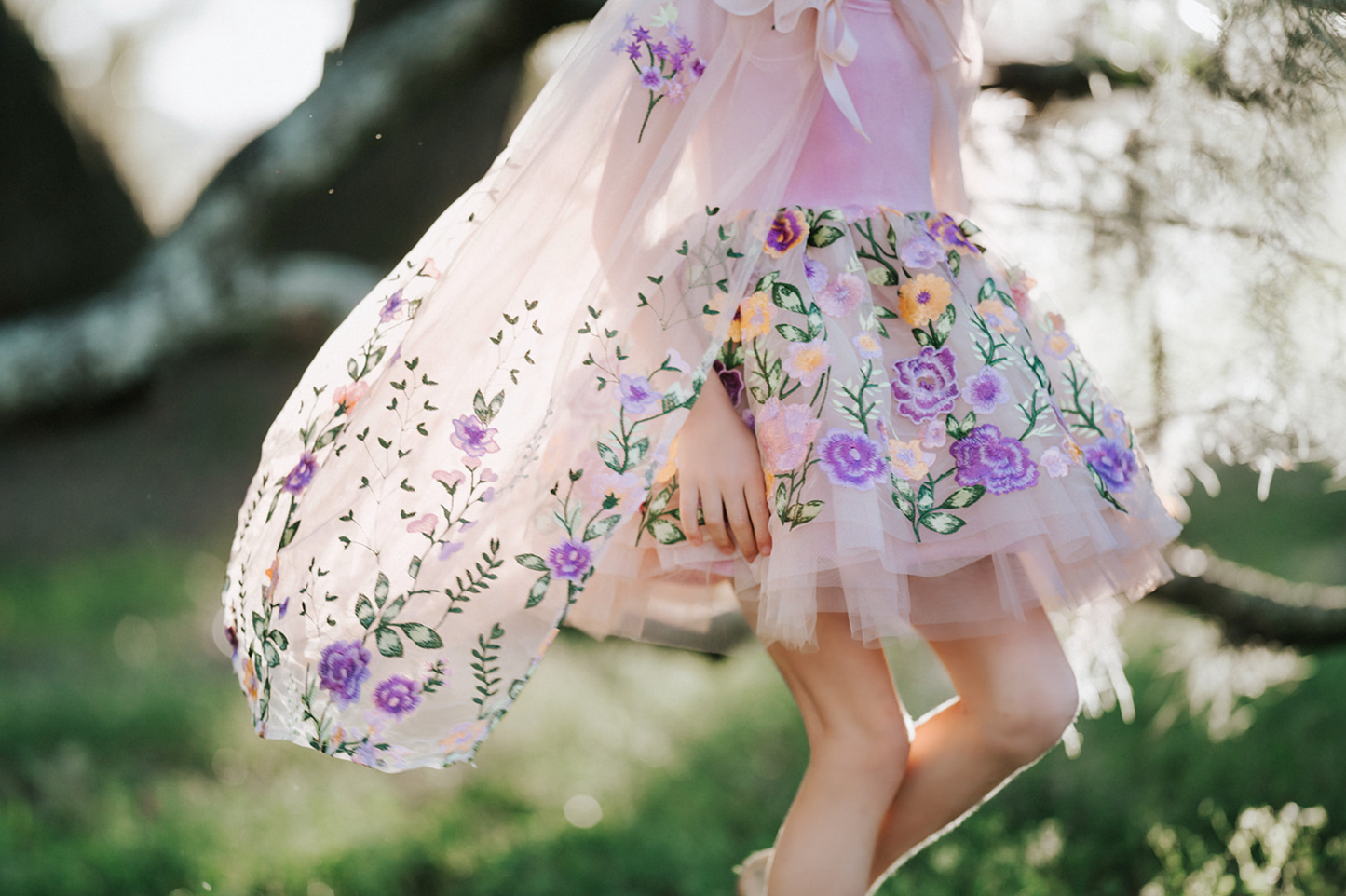 A young girl is dressed in a spring floral tutu dress. Lavender velvet bodice with short sleeves and fluffly layers of ivory tulle. The skirt is embroidered with peach, purple, pink flowers and green vines. A matching ivory cape with floral pattern is tied around her shoulders.