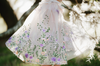 A young girl is dressed in a spring floral tutu dress. Lavender velvet bodice with short sleeves and fluffly layers of ivory tulle. The skirt is embroidered with peach, purple, pink flowers and green vines. A matching ivory cape with floral pattern is tied around her shoulders.