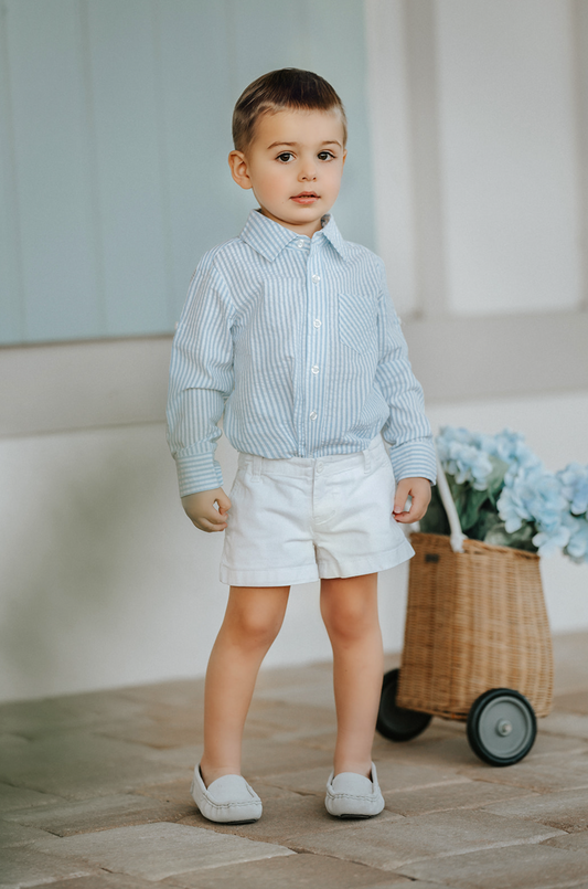 A young boy wears a long sleeve button up collared shirt. A light blue and white vertical seersucker shirt with a pocket on the left chest.