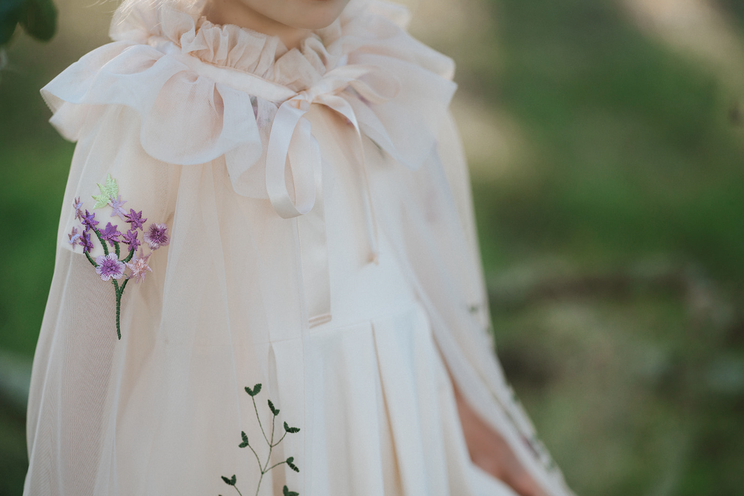 A young girl is dressed in a spring floral tutu dress. Lavender velvet bodice with short sleeves and fluffly layers of ivory tulle. The skirt is embroidered with peach, purple, pink flowers and green vines. A matching ivory cape with floral pattern is tied around her shoulders.