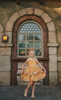 A young girl with blonde hair stands outdoors, wearing the Portrait Petite Red Rose Princess twirl length gown, an intricately designed gold princess dress adorned with floral lace patterns. The satin off shoulder neckline is adorned with a gold jewel.