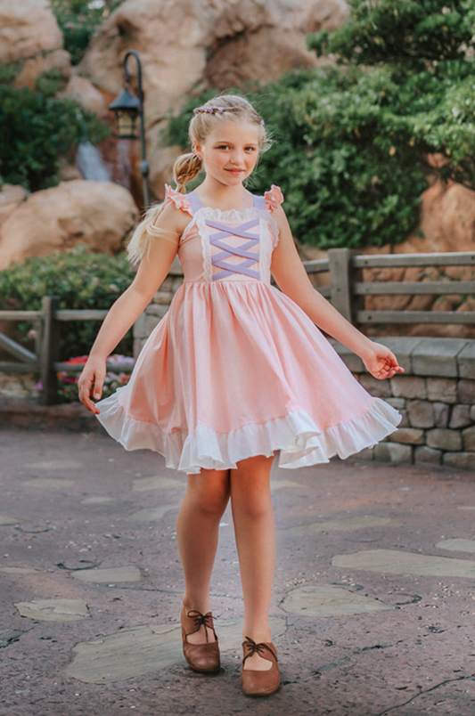 A young girl wearing Our Original Rapunzel Twirl Dress with a lavender laced bodice and white frills twirls outdoors. The dress is a light pink with white ruffle at the hem. The sleeveless dress has light pink ruffles on lavender straps.