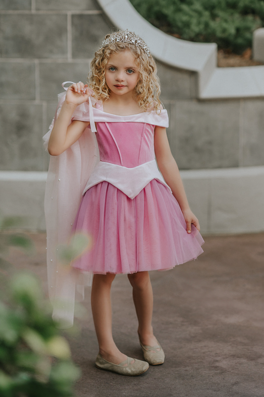 A young girl in an exquisite Portrait Petite Sleeping Beauty twirl length gown poses in a castle setting. The pink stretch velvet gown has an off the shoulder neckline with two straps. The pink tulle skirt is full and has a cotton liner for comfort.